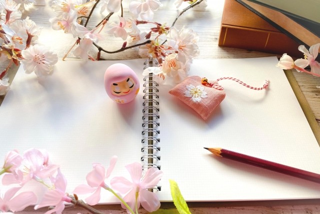 白いノートの上に桜の花と鉛筆とピンクの達磨が置いてある